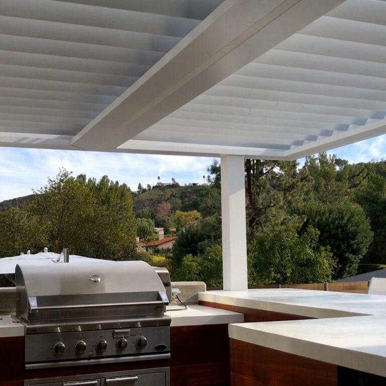 Under-canopy view of a white aluminum louvered pergola with open louvers over an outdoor kitchen featuring a stainless steel built-in grill and white quartz countertop, with a wooded hillside and canyon view beyond