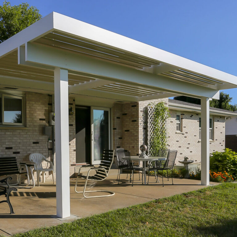 White aluminum louvered pergola with open louvers attached to a single-story painted brick ranch home, covering a concrete patio with casual seating, a small dining set, and flowering garden beds