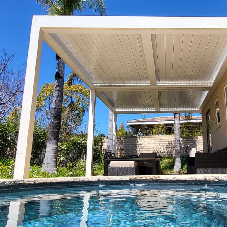 Low-angle view of a white aluminum louvered pergola with closed louvers over a pool patio surrounded by palm trees under a deep blue sky