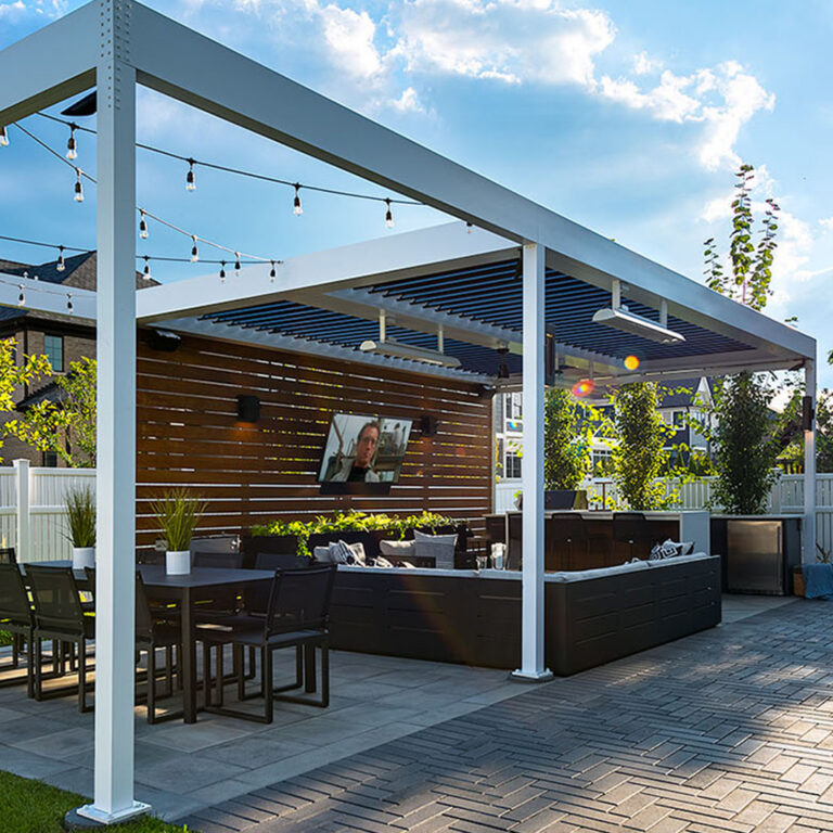 White aluminum louvered pergola with open louvers and string lights at dusk over a paver patio with a dining table, lounge seating, outdoor TV, ceiling fan, infrared heaters, and a wood-clad feature wall