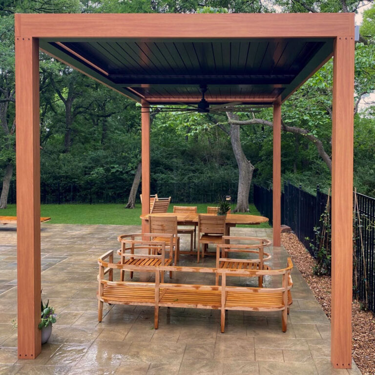 Freestanding aluminum louvered pergola with warm wood-grain finish and fully closed dark louvers over a teak circular bench set and oval dining table on a flagstone patio with a wooded backyard