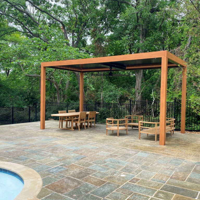 Freestanding aluminum louvered pergola with a warm wood-grain finish and ceiling fan over a teak dining and lounge set on a flagstone pool patio, framed by mature oak trees