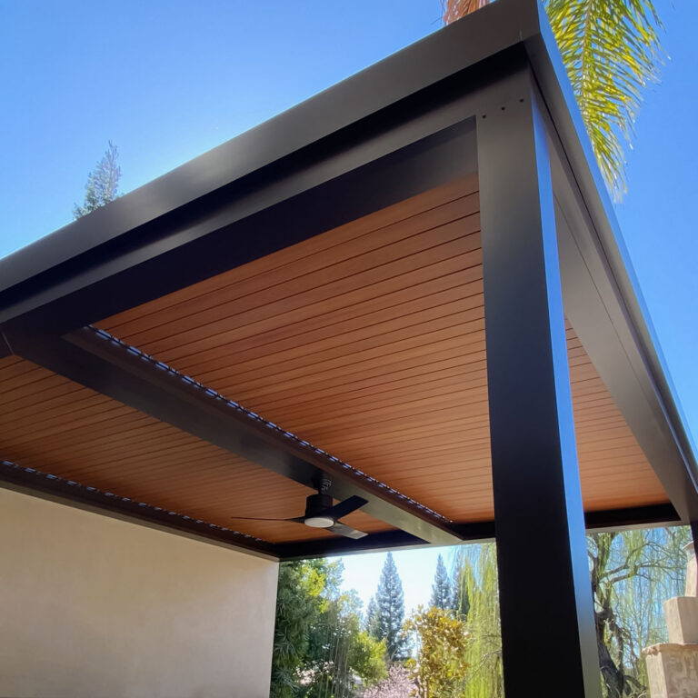 Underside of a heavy-duty extruded aluminum pergola with closed woodgrain louvers, stepped fascia detail, and a ceiling fan mounted to the beam