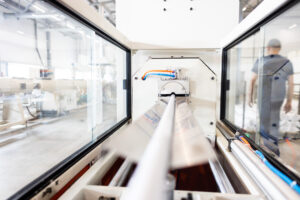 Close-up view through the glass enclosure of an industrial aluminum extrusion machine as a profile exits the die, with a factory worker visible in the background