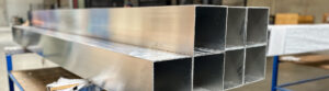 Stacked raw aluminum square tube extrusions on a workbench inside a pergola manufacturing facility
