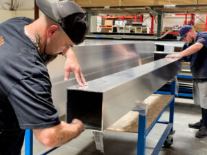 Two factory workers inspecting and handling a raw aluminum pergola post on a production workbench, with shelving and components visible behind them