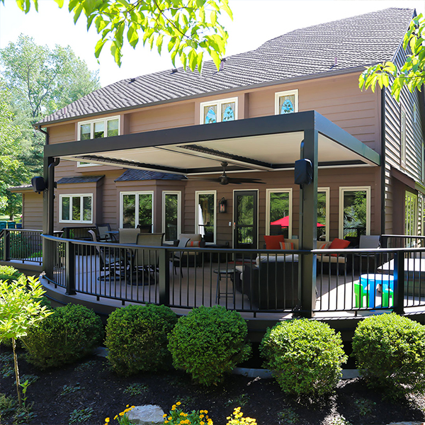 A closed louvered roof pergola with an integrated ceiling fan covers a curved backyard deck furnished with lounge seating.