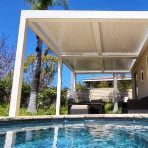 White aluminum louvered pergola with louvers closed, shading a poolside seating area next to palm trees