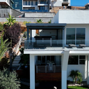 A gray aluminum pergola frames an upper-level terrace with glass railing, overlooking a modern hillside neighborhood.