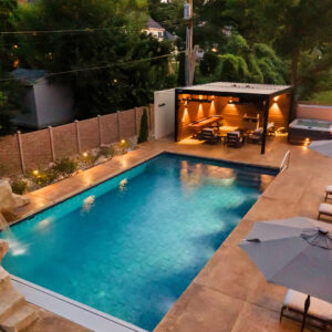 Black louvered pergola over a wood-paneled cabana with wall sconces, dining table, and hot tub beside a lit backyard pool at dusk
