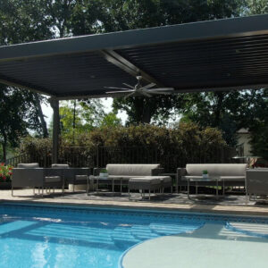 Aluminum louvered pergola with closed roof and mounted ceiling fan over a cushioned lounge set beside a curved pool