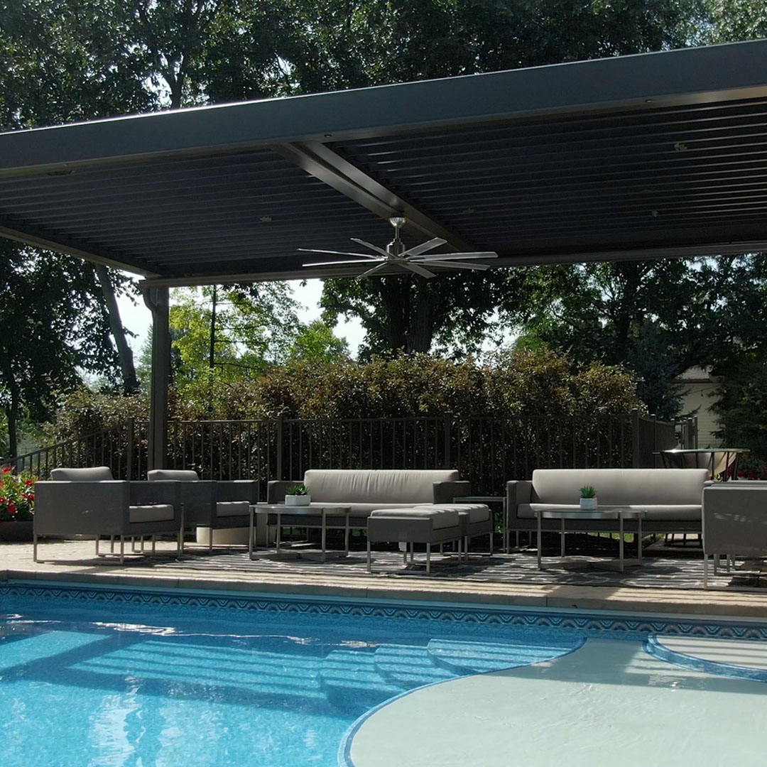 Aluminum louvered pergola with closed roof and mounted ceiling fan over a cushioned lounge set beside a curved pool