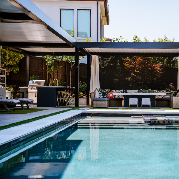 Dark aluminum louvered pergola sections shading an outdoor kitchen with grill and a lounge seating area beside a pool
