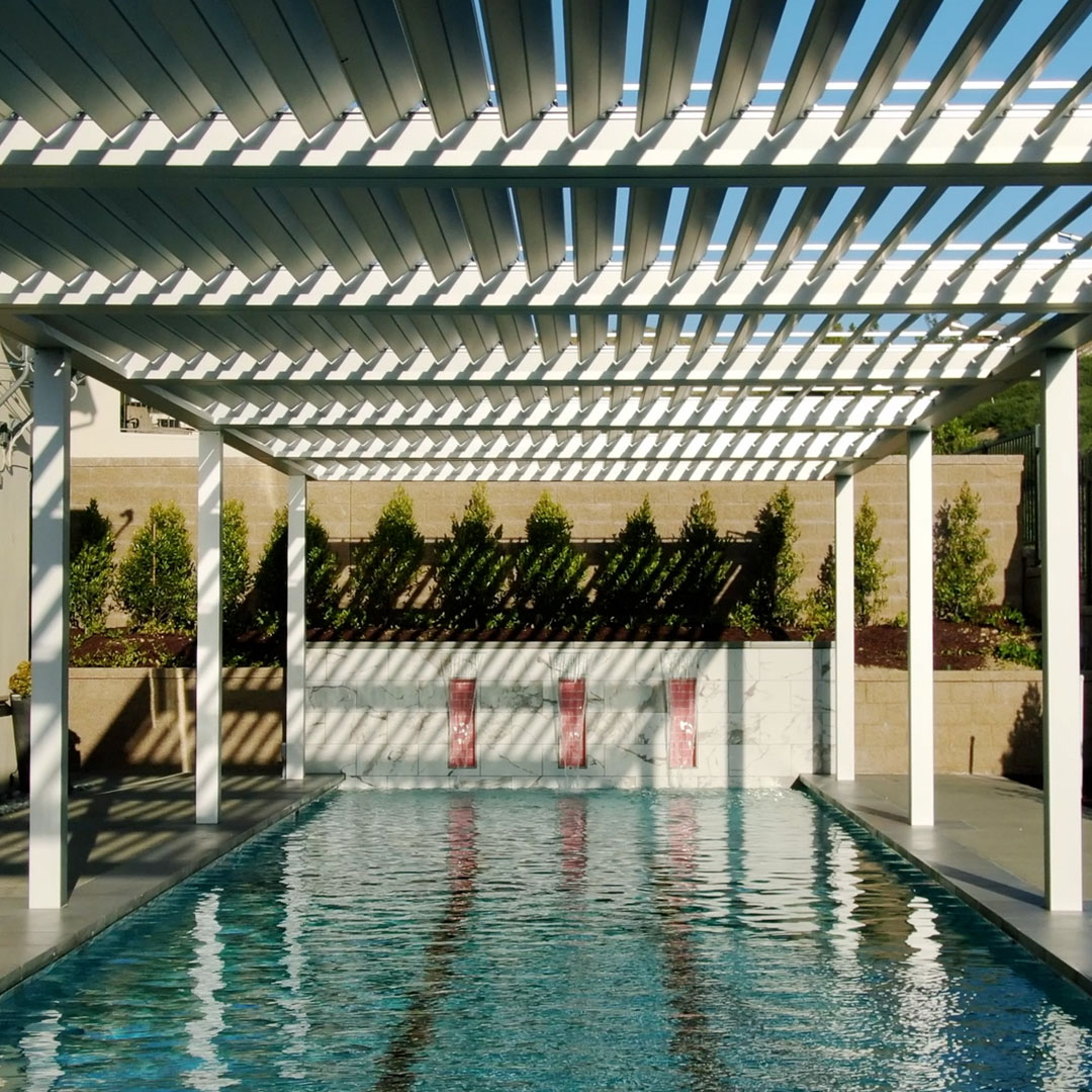 White aluminum louvered pergola with louvers open to blue sky, casting striped shadows across a lap pool and water wall