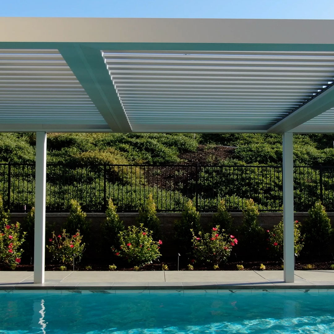 White aluminum pergola with louvers open at an angle above a pool deck, flower beds, and iron fence line